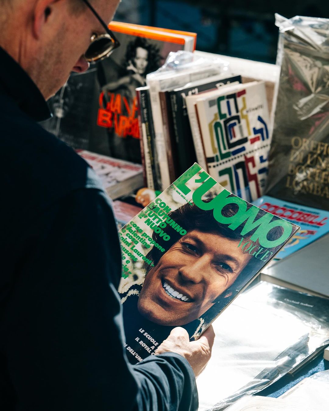 Adult male examines 'L'Uomo Vogue' magazine with a smiling man on the cover, surrounded by a magazine stand outdoors.