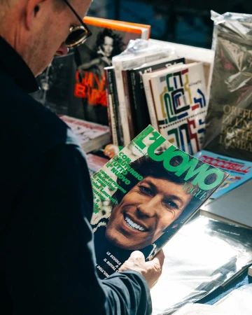 Adult male examines 'L'Uomo Vogue' magazine with a smiling man on the cover, surrounded by a magazine stand outdoors.