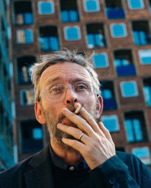 Adult male smoking a cigarette in an urban setting, wearing glasses and a dark jacket, with a multi-story building in the background.
