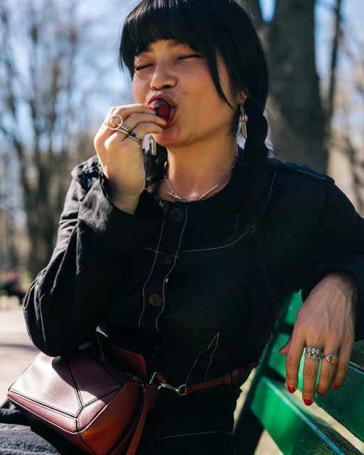 Asian woman with dark hair in braids eats a red fruit outdoors. She wears a black jacket, jewelry, and a maroon cross-body purse.