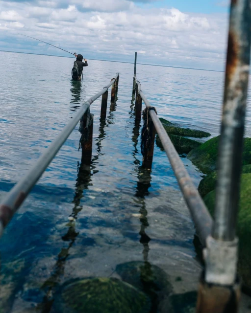 Angler wading in the ocean, casting a fishing rod near a weathered pier, under a partly cloudy sky, captured in a medium shot.