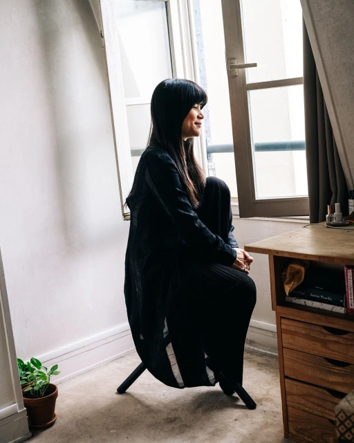 Woman in black outfit seated by window in sunlit room. Minimalist decor, featuring wooden desk, potted plant, and natural light.