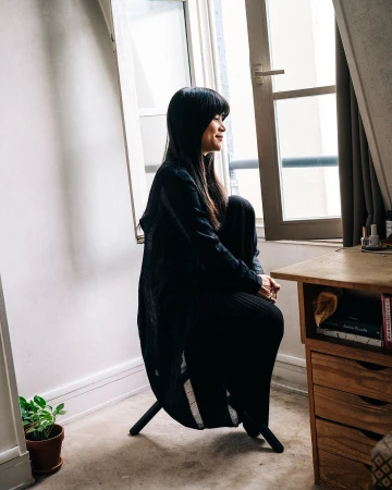 Woman in black outfit seated by window in sunlit room. Minimalist decor, featuring wooden desk, potted plant, and natural light.