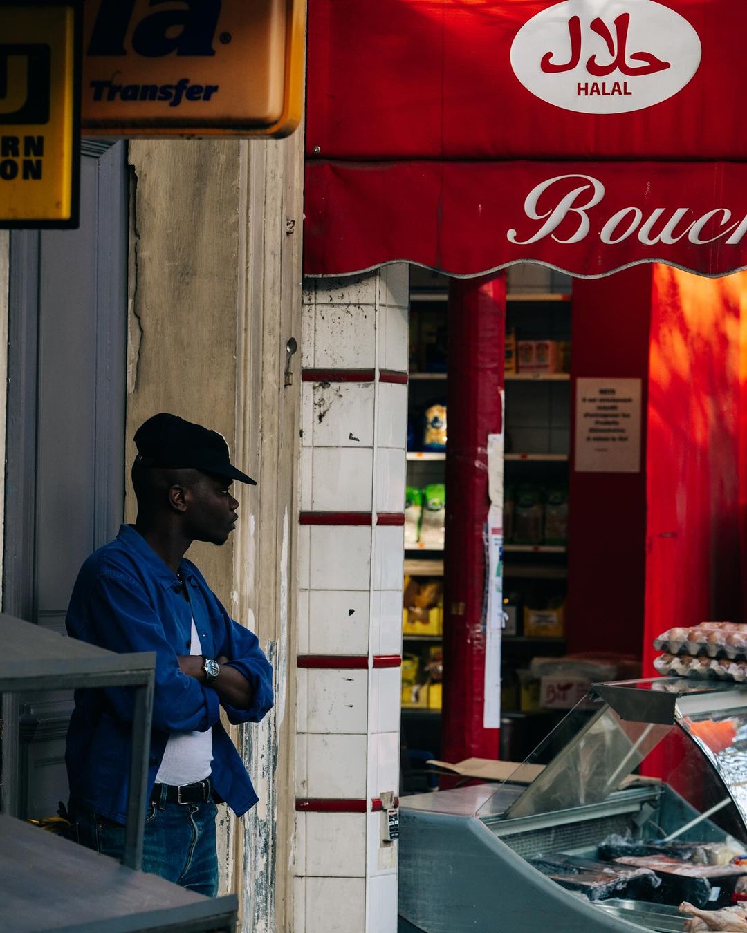Adult Black man in blue work jacket with crossed arms stands outside a butcher shop with 'Halal' signage in an urban setting.