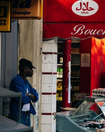Adult Black man in blue work jacket with crossed arms stands outside a butcher shop with 'Halal' signage in an urban setting.