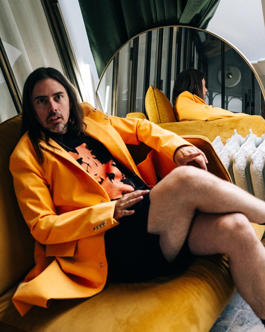 Man with long brown hair wearing a bright yellow blazer seated on a yellow velvet sofa in New York by Adam Katz Sinding