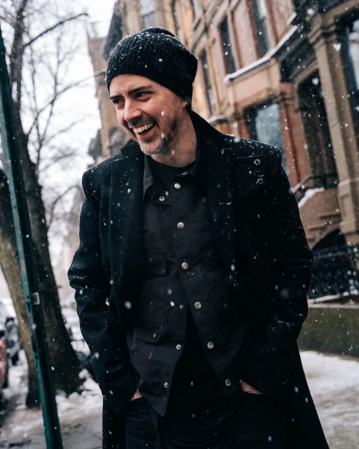 Man in black coat smiles outdoors in winter snowfall near brownstone buildings, wearing a beanie and button-down shirt.