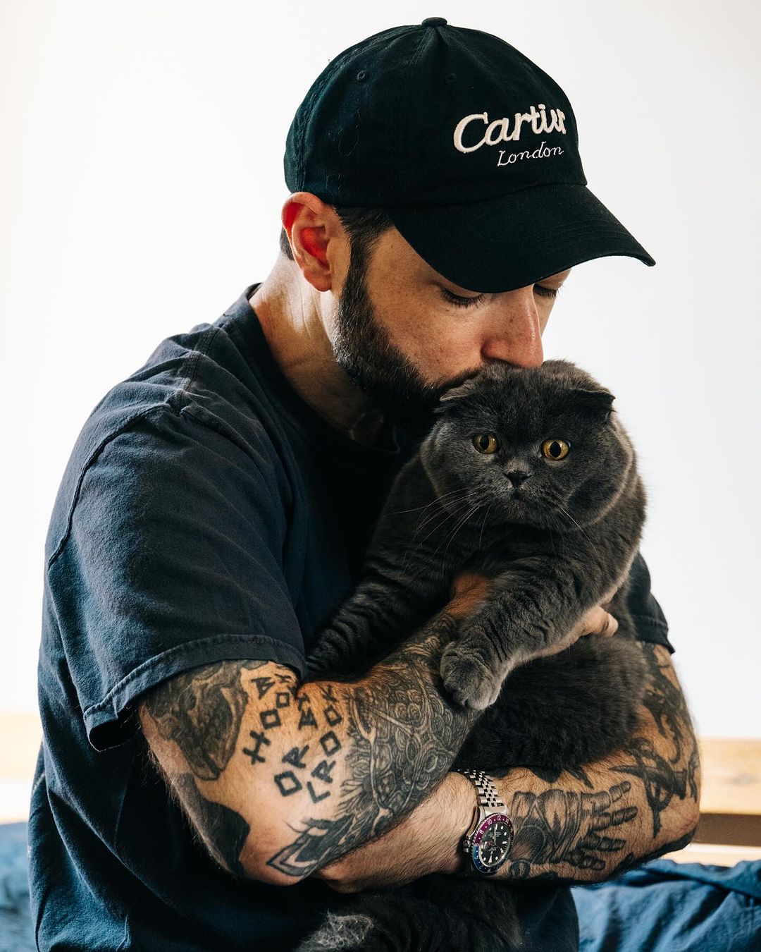 Bearded man wearing a black baseball cap kissing a fluffy grey cat while holding it in his arms