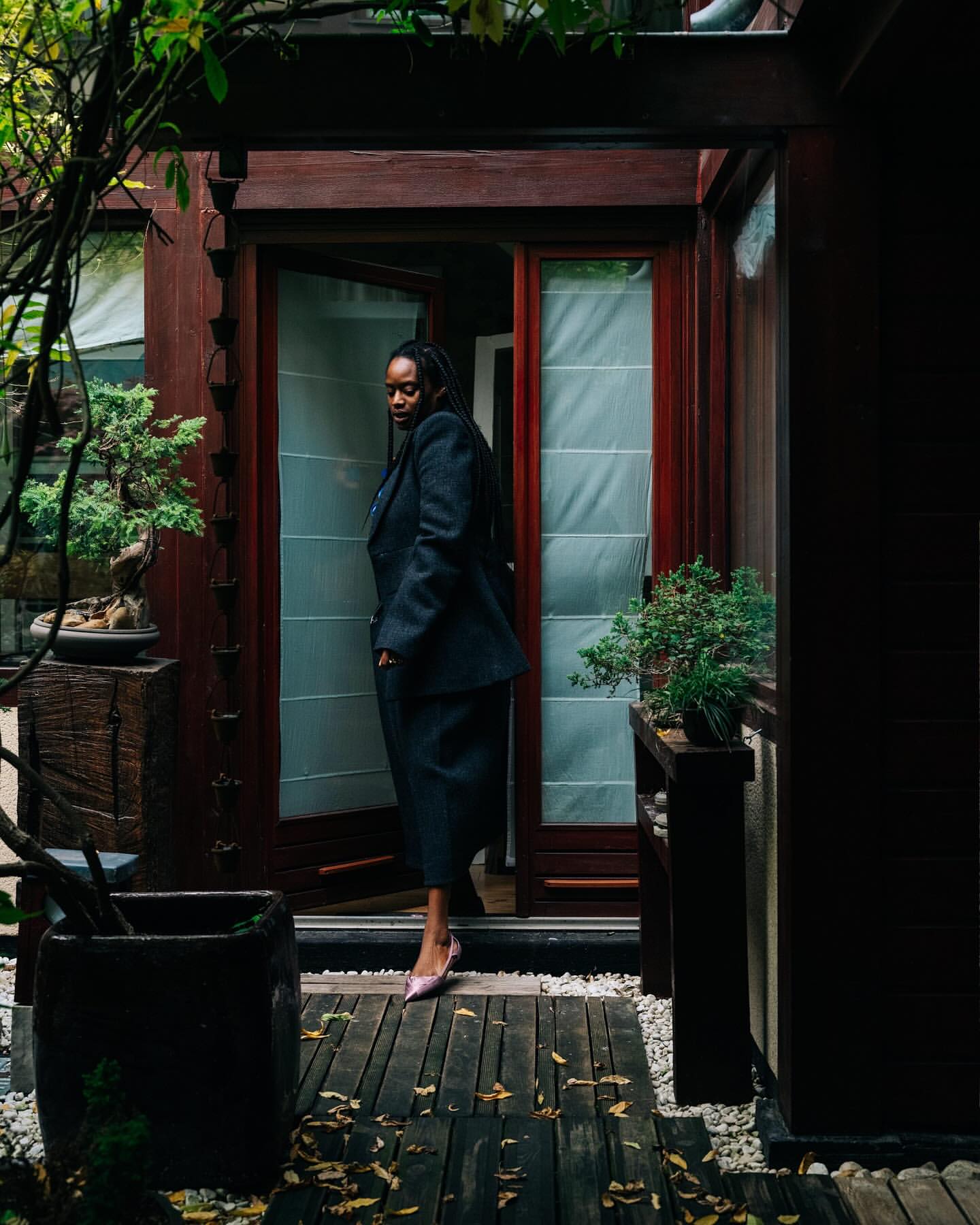 A stylish Black woman in a blazer and skirt poses in front of a doorway, surrounded by greenery, potted plants, and a wooden walkway.