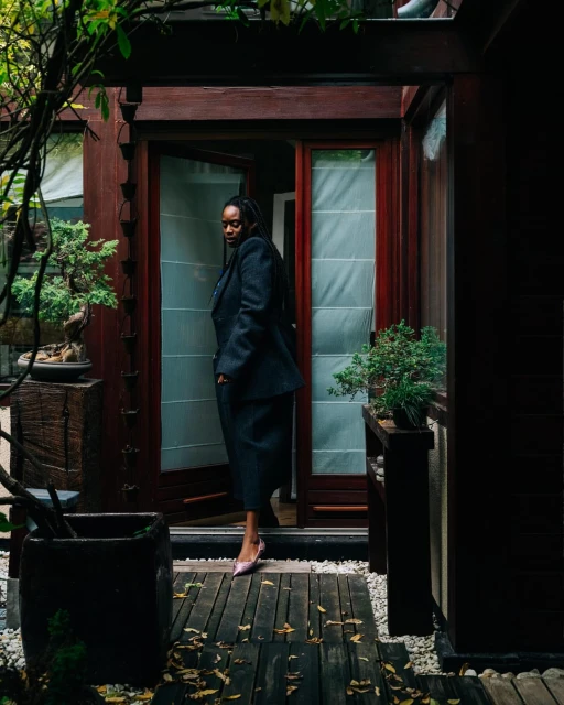 A stylish Black woman in a blazer and skirt poses in front of a doorway, surrounded by greenery, potted plants, and a wooden walkway.