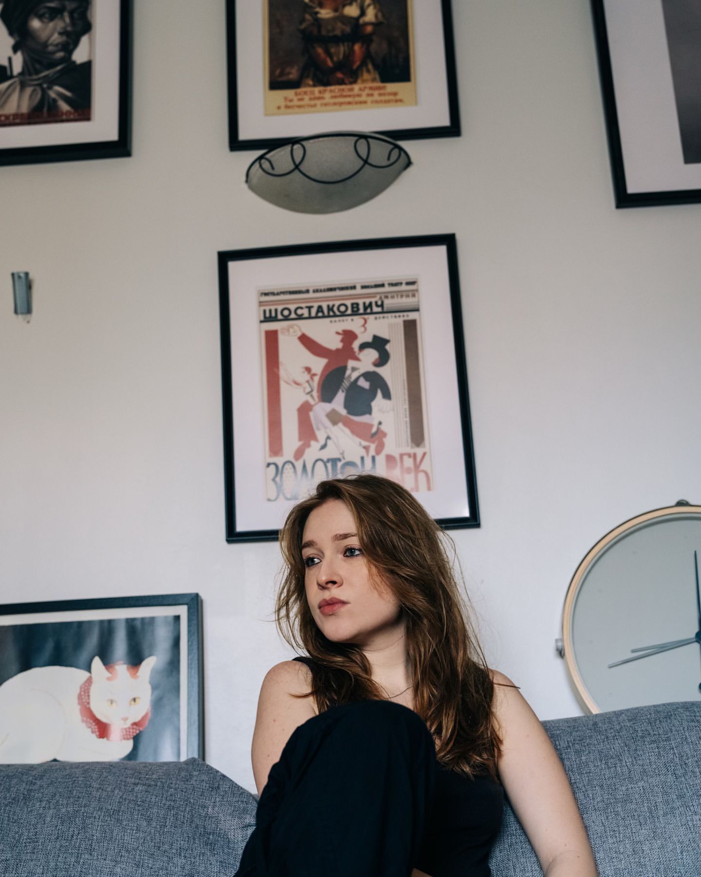 Indoor portrait of a young adult woman with long brown hair sitting on a gray couch against a white wall with framed artwork.