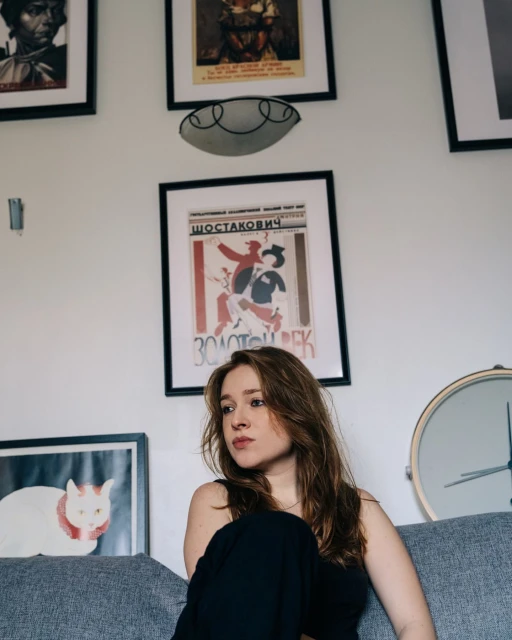 Indoor portrait of a young adult woman with long brown hair sitting on a gray couch against a white wall with framed artwork.