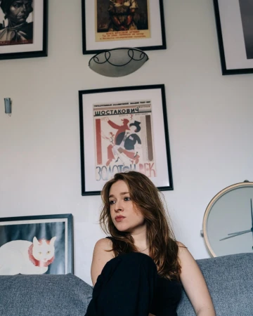 Indoor portrait of a young adult woman with long brown hair sitting on a gray couch against a white wall with framed artwork.