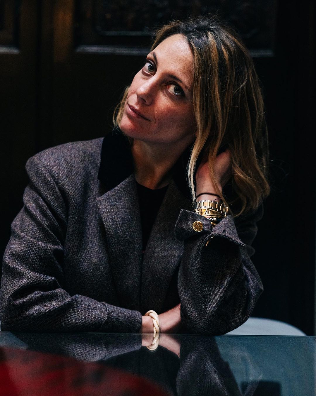 Portrait of a woman with hand near her head sitting at a shiny table in a dark indoor environment wearing a grey jacket.