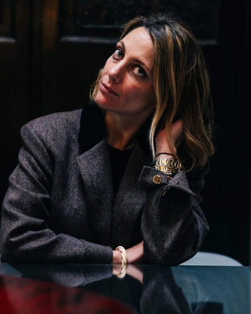 Portrait of a woman with hand near her head sitting at a shiny table in a dark indoor environment wearing a grey jacket.