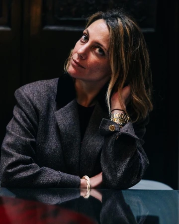Portrait of a woman with hand near her head sitting at a shiny table in a dark indoor environment wearing a grey jacket.