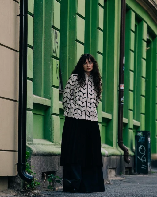 A stylish woman stands in front of a green wall wearing a cream and black patterned jacket and long black skirt on a city street.