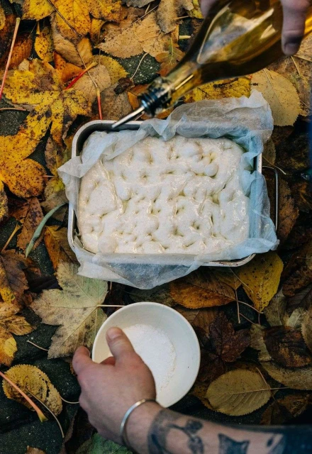 Preparing focaccia dough: person pouring olive oil on the dough in a baking tin surrounded by autumn leaves on the ground.