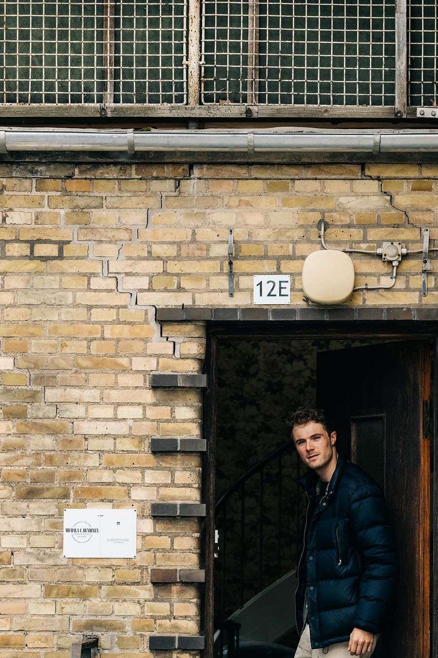 Young adult male stands in doorway of brick building with window and sign during daytime, looking at camera with neutral expression.