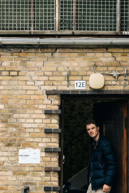 Young adult male stands in doorway of brick building with window and sign during daytime, looking at camera with neutral expression.
