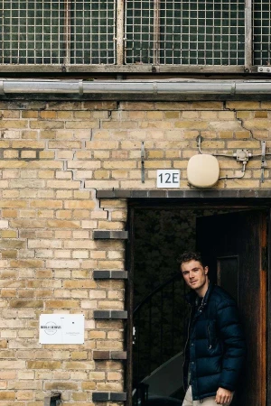 Young adult male stands in doorway of brick building with window and sign during daytime, looking at camera with neutral expression.