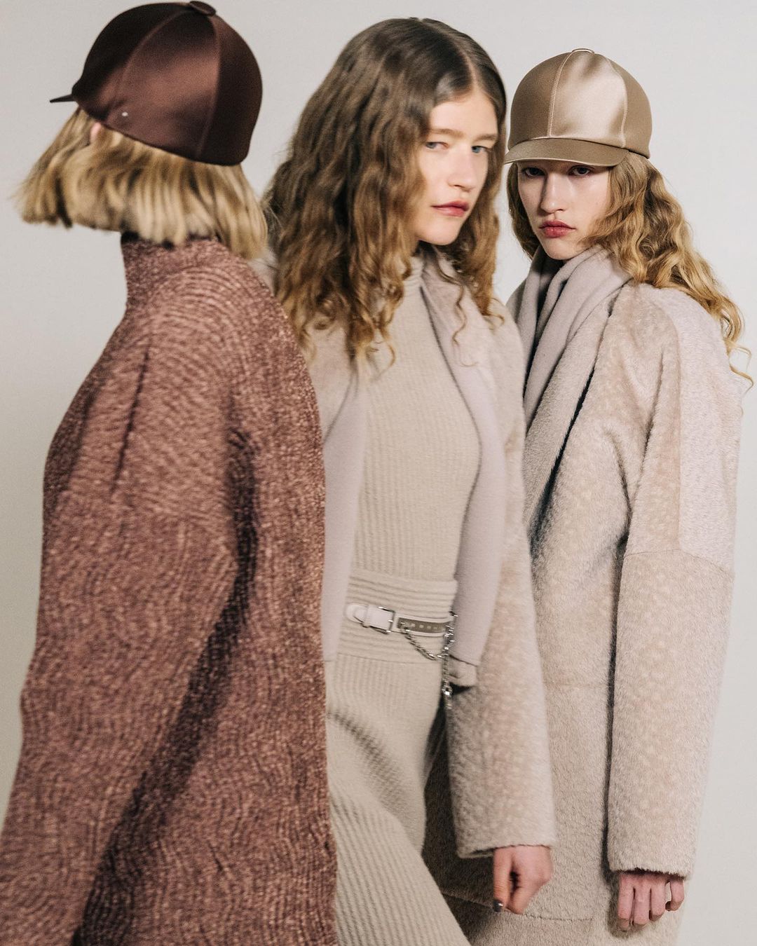 Three fashion models pose indoors, wearing stylish hats and neutral-toned knitwear, featuring soft textures and a minimalist aesthetic.