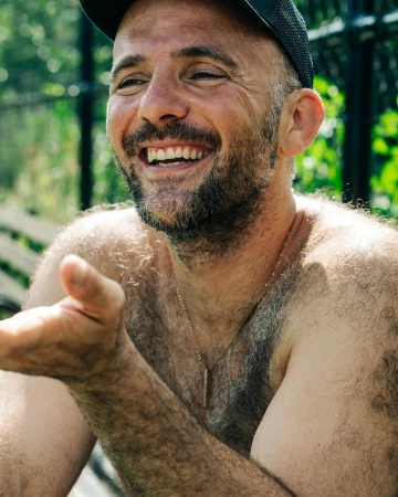 Close-up portrait of a smiling shirtless man wearing a baseball cap with his hand raised, photographed in New York.