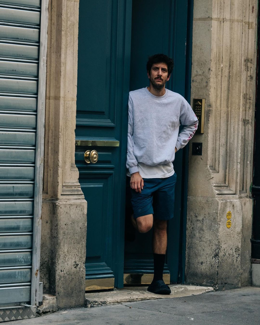 Man standing in a doorway wearing a light grey crewneck sweatshirt, blue shorts, and dark shoes
