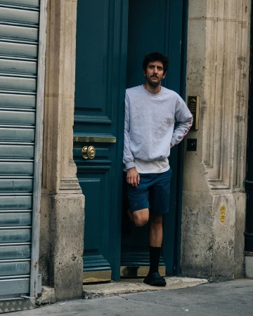 Man standing in a doorway wearing a light grey crewneck sweatshirt, blue shorts, and dark shoes