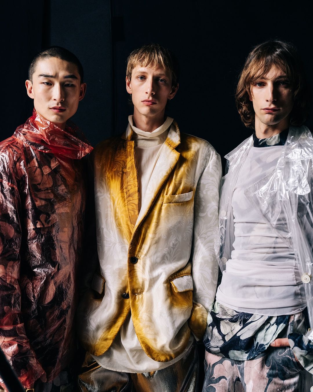 Fashion photo of three young adult men in translucent floral patterned jackets posing in a studio with dark background and eye-level camera angle.