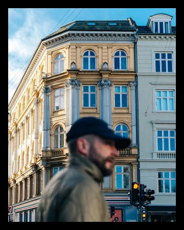 A blurred man in a dark cap and jacket walks past ornate classical buildings in New York captured by Adam Katz Sinding.