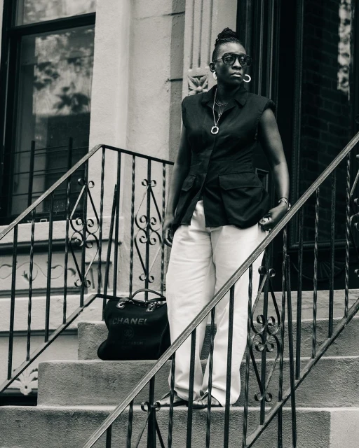 Monochrome outdoor shot of an adult woman in sunglasses posing on stairs with a Chanel tote bag next to her, city building backdrop.