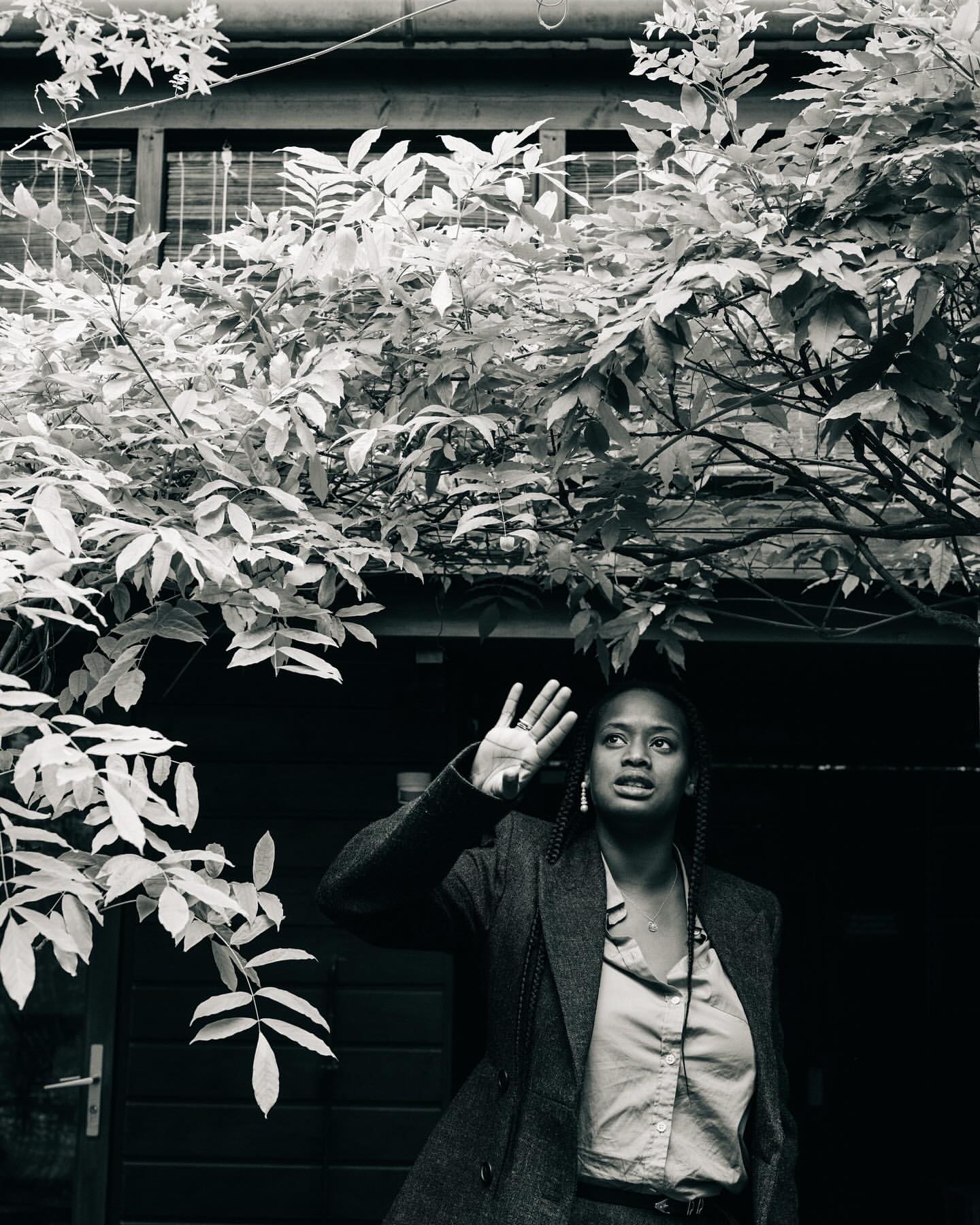 Monochrome portrait of a Black adult female in a suit jacket raising her hand outdoors, with leafy trees overhead and a building behind.