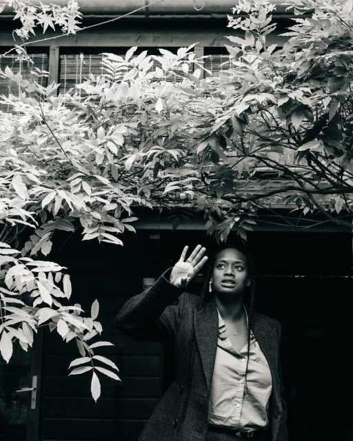 Monochrome portrait of a Black adult female in a suit jacket raising her hand outdoors, with leafy trees overhead and a building behind.