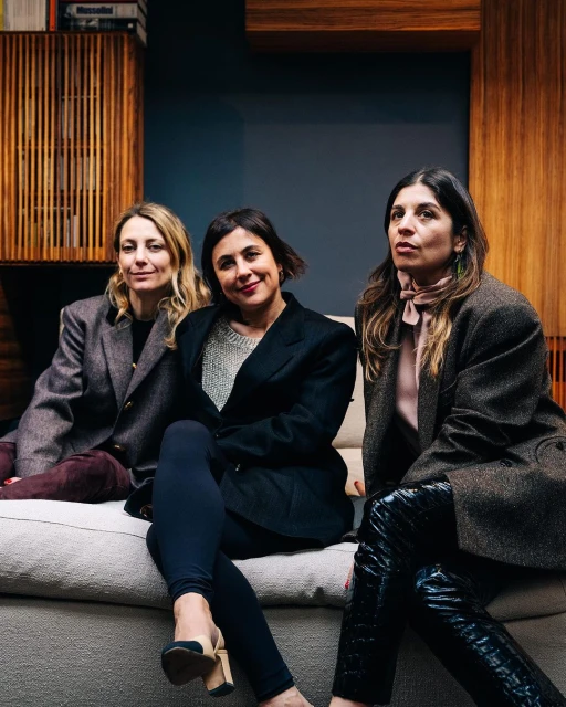 Three adult women are sitting on a sofa in an indoor setting, posing for a medium shot in blazers and fashionable pants, with wood paneling in background.