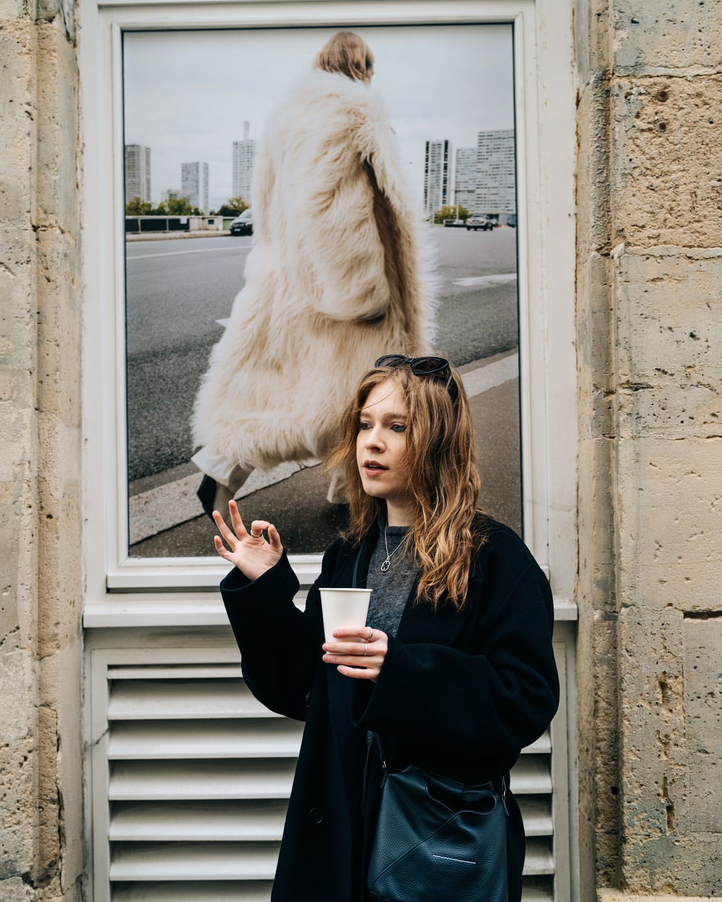 Adult woman wearing a black coat and sunglasses on her head, holding a cup, posing near a framed photograph of a model wearing a coat.
