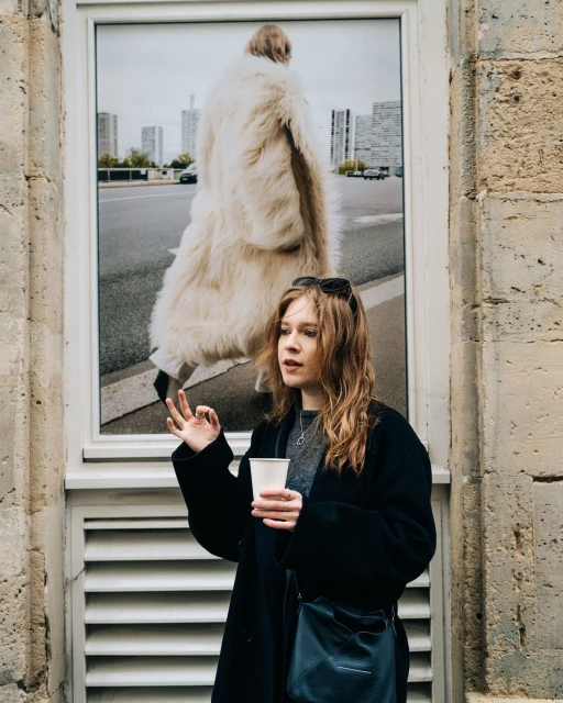 Adult woman wearing a black coat and sunglasses on her head, holding a cup, posing near a framed photograph of a model wearing a coat.