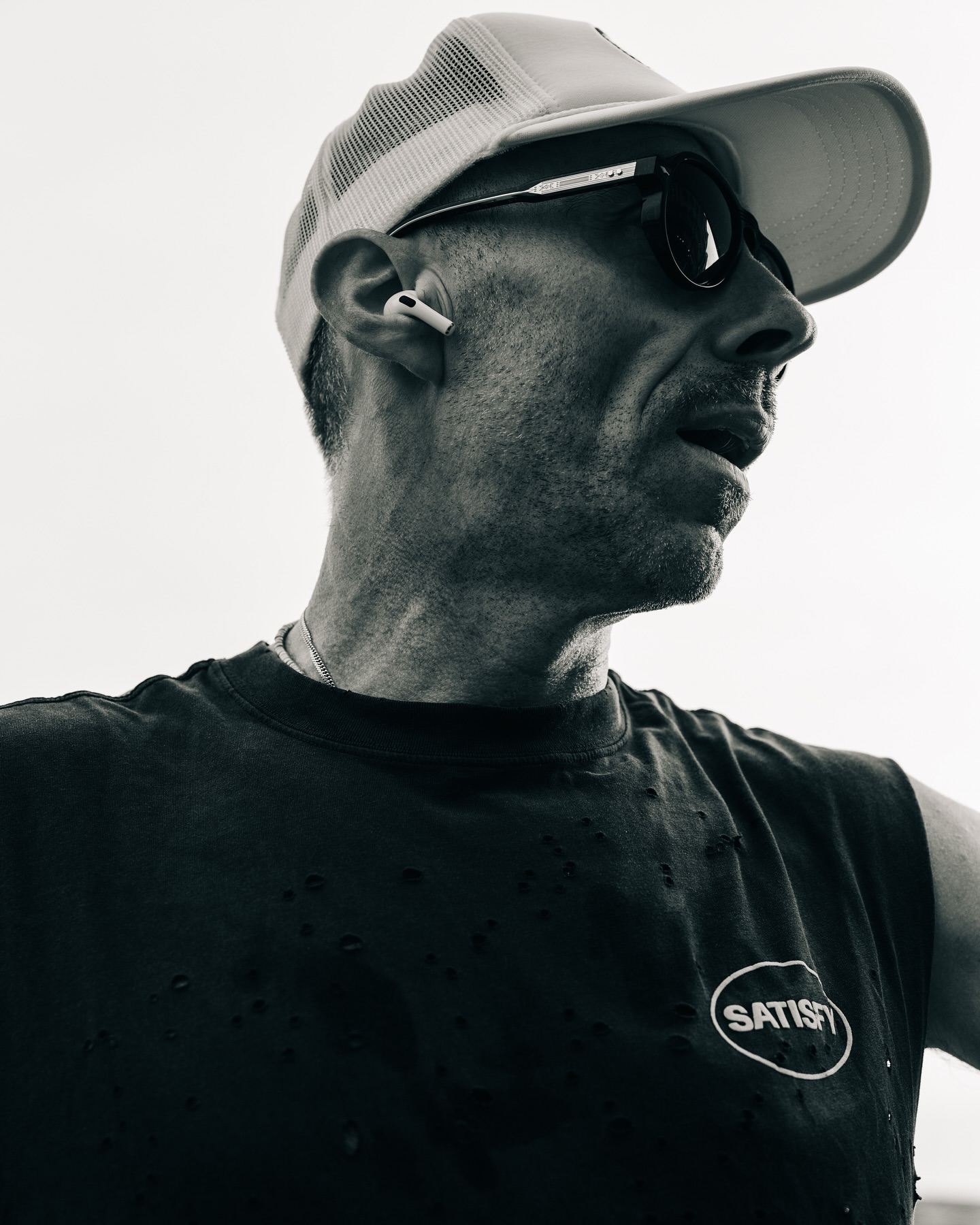 Monochrome portrait of an adult man wearing a cap and sunglasses, looking off to the side, with a bright, neutral background.