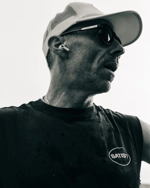 Monochrome portrait of an adult man wearing a cap and sunglasses, looking off to the side, with a bright, neutral background.