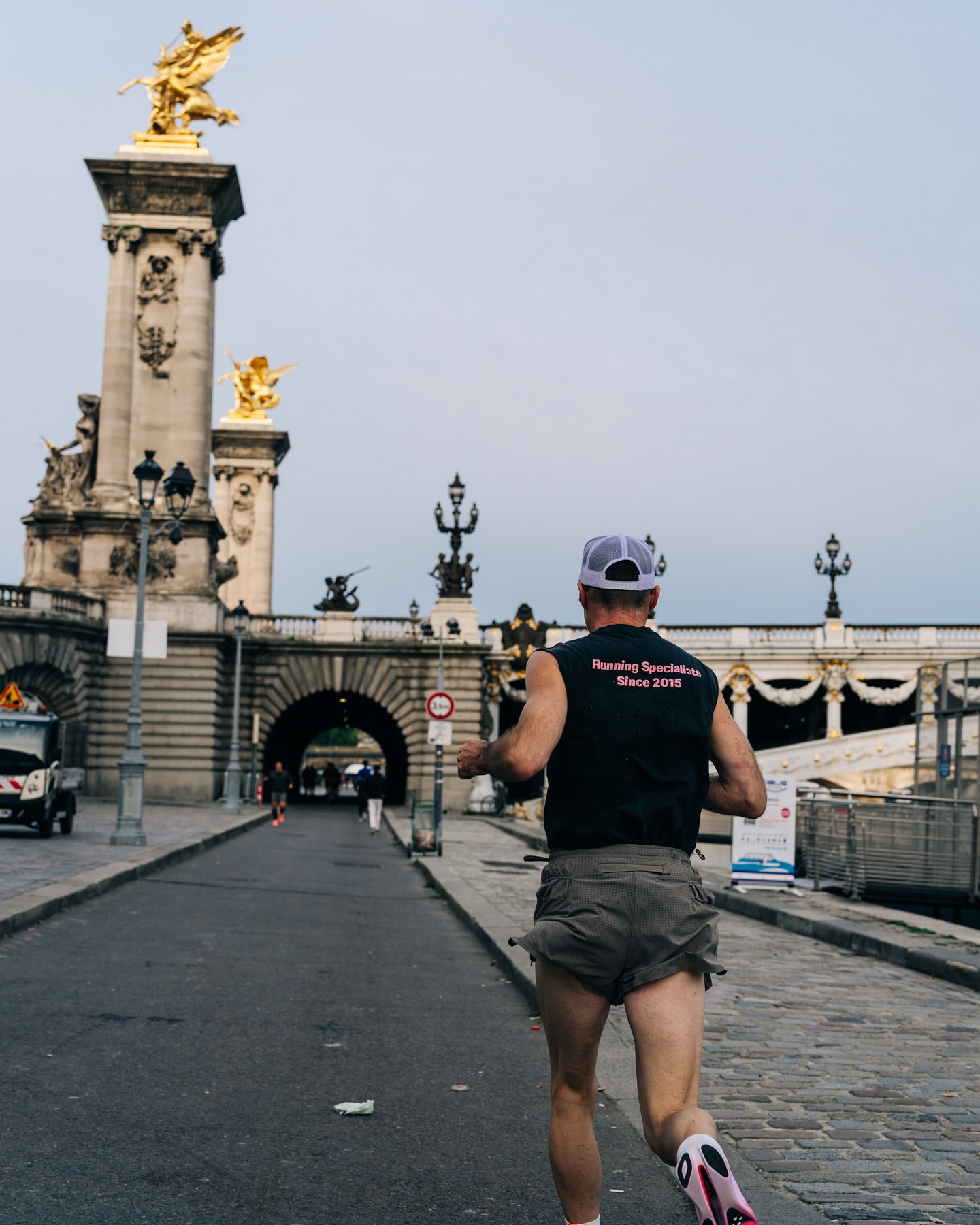Adult male in running attire jogs along stone street in Paris, France, with ornate golden statues on bridges under overcast sky.