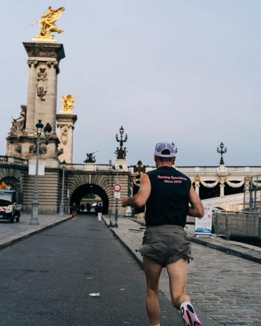 Adult male in running attire jogs along stone street in Paris, France, with ornate golden statues on bridges under overcast sky.