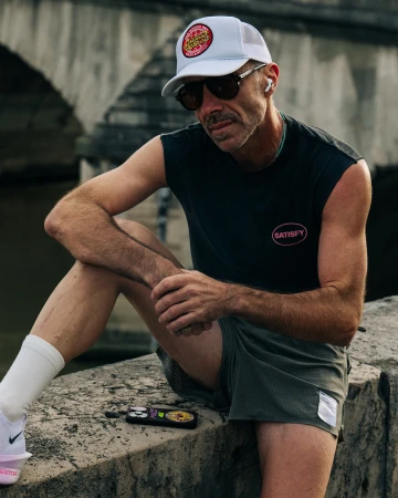 Man wearing a white baseball cap, sunglasses, and black sleeveless shirt seated outdoors by a stone bridge.