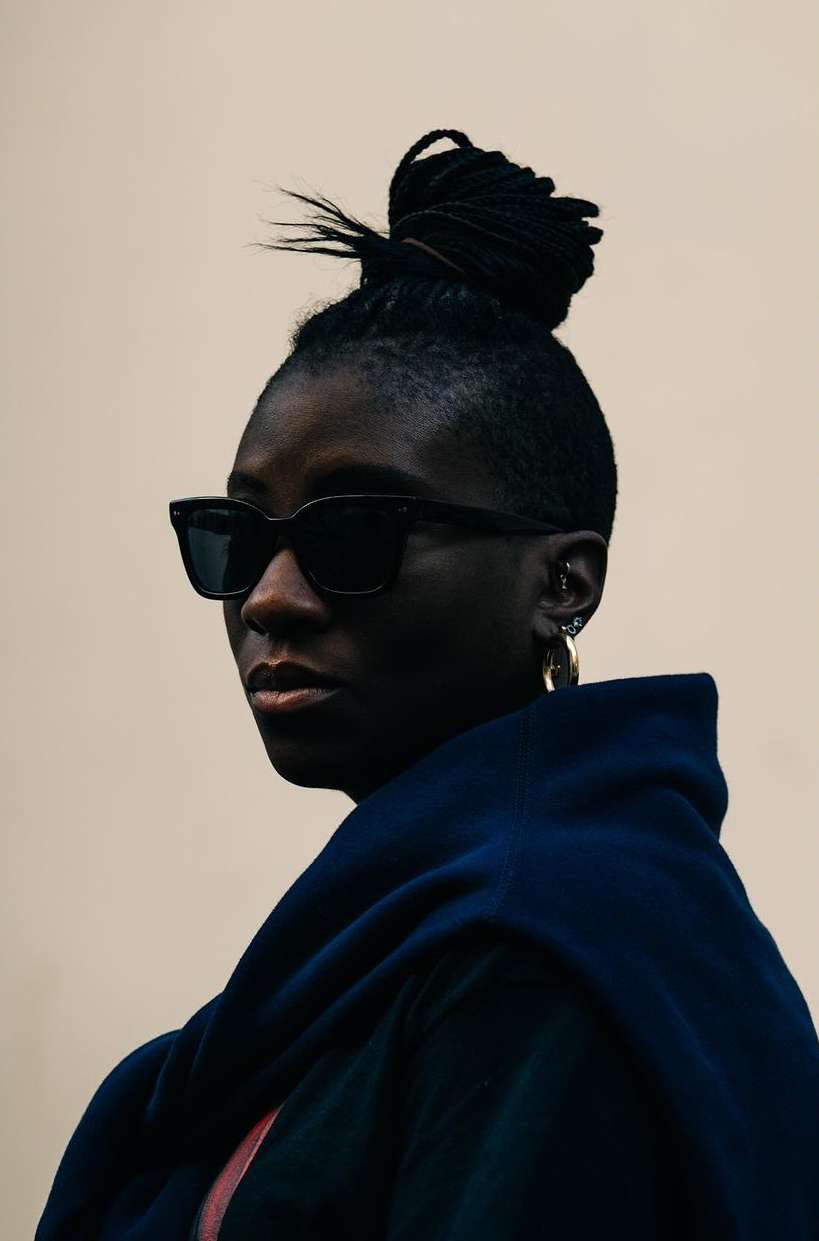 Portrait of a stylish adult female with braided hair posing indoors in sunglasses and a blue hooded jacket against a neutral wall.