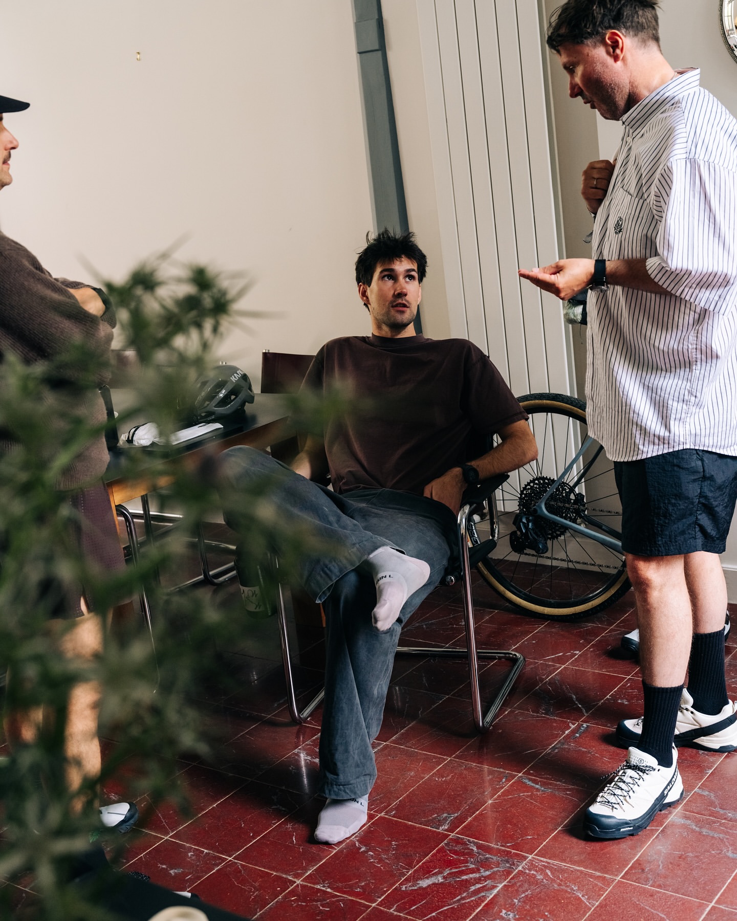 Three adult men casually converse indoors near a bicycle and houseplant, wearing casual attire under indoor lighting, creating an informal setting.
