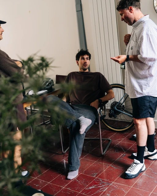 Three adult men casually converse indoors near a bicycle and houseplant, wearing casual attire under indoor lighting, creating an informal setting.