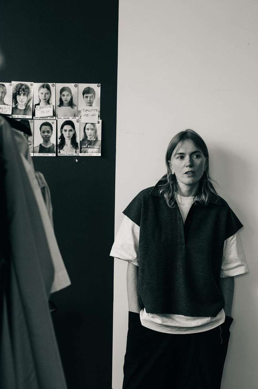 Monochrome portrait of a woman standing indoors against a two-tone wall with model polaroids pinned up behind her.