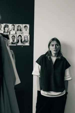 Monochrome portrait of a woman standing indoors against a two-tone wall with model polaroids pinned up behind her.