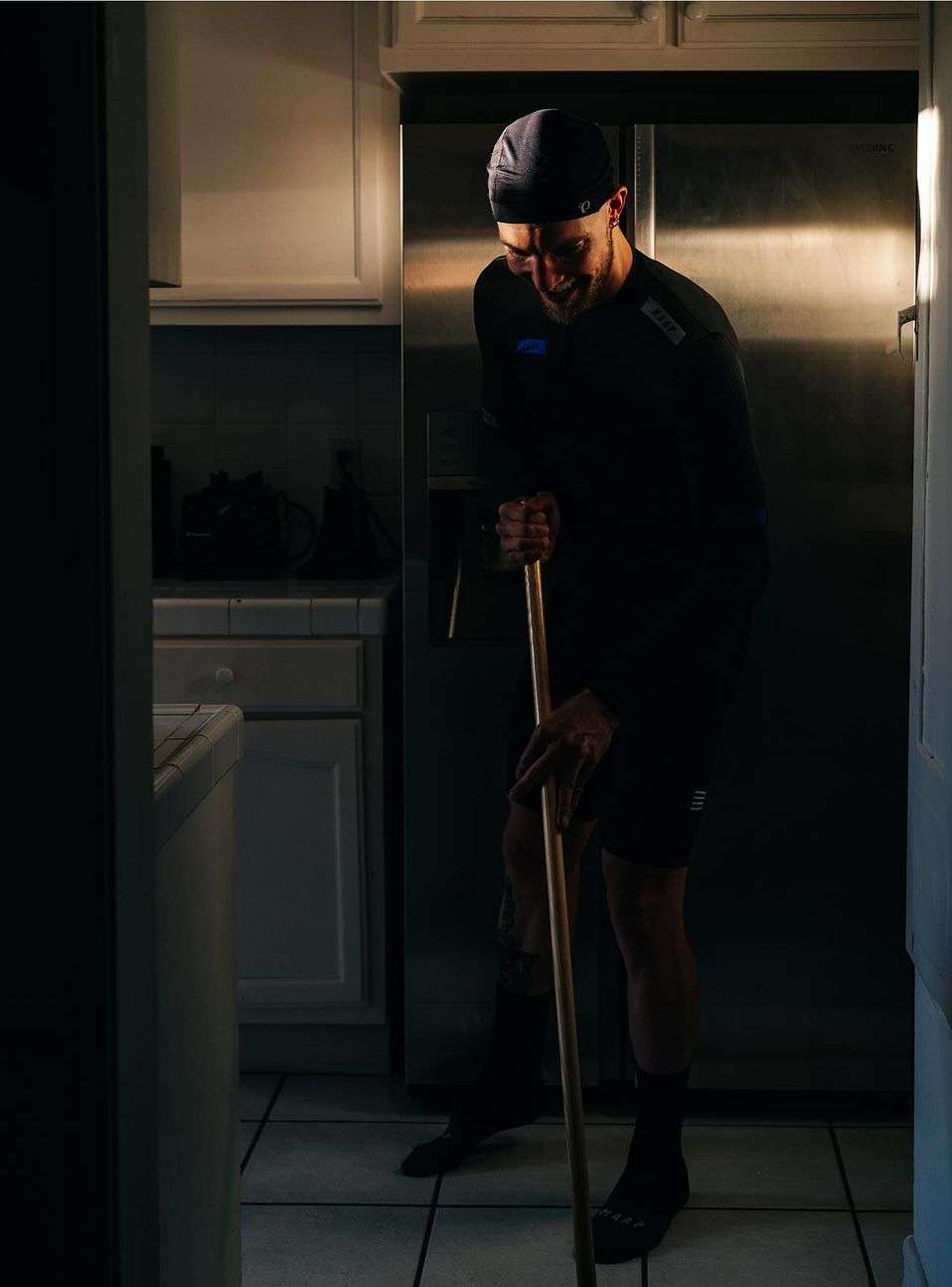 Adult man with a slight smile leans forward while holding a mop handle in a kitchen with tile flooring and stainless steel appliances.