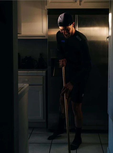Adult man with a slight smile leans forward while holding a mop handle in a kitchen with tile flooring and stainless steel appliances.