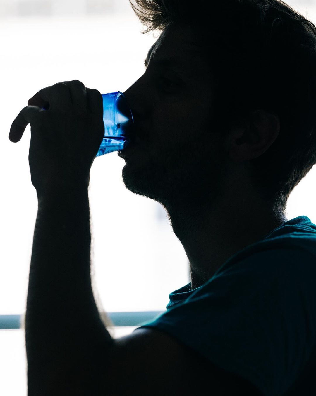 Silhouette of a man drinking water from a blue glass, side profile, bright window background, indoor setting, health and wellness.
